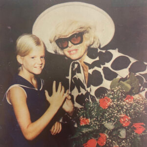 Carol Channing wearing a big hat and sunglasses while holding roses and posing with a young girl