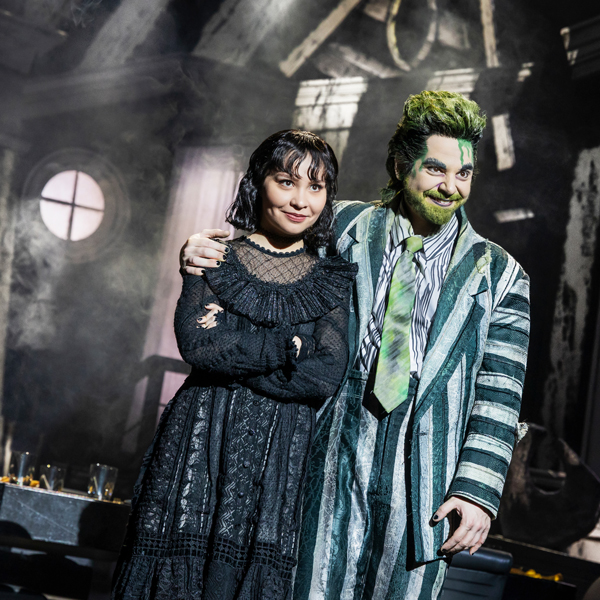 A girl wearing an old-fashioned black dress and a man in a striped suit with green hair and beard smile menacingly.