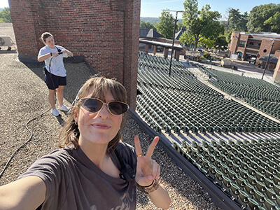 A woman wearing sunglasses smiles and makes a peace sign while taking a selfie from a rooftop overlooking an outdoor amphitheater filled with empty green seats. Another person in the background holds a camera or similar equipment. The scene is bright and sunny, with trees and brick buildings visible in the distance.