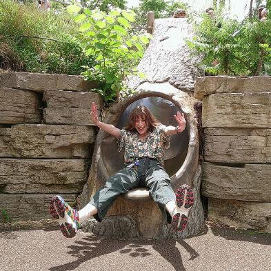 A person with shoulder-length brown hair is joyfully sliding down a playground tunnel slide made to look like a hollowed-out tree trunk. They are wearing a patterned shirt, green pants, and colorful shoes with red soles. Their arms are raised in excitement as they come out of the slide, surrounded by large stacked stones and lush greenery.
