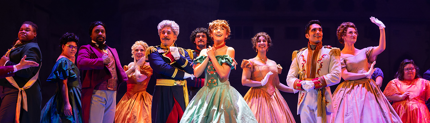 A lively ensemble cast performs a grand ballroom scene from Disney's Frozen on stage. At the center, Anna, dressed in a green and gold gown with floral accents, clasps her hands with excitement. To her right, Hans stands confidently in a white military-style jacket with gold details. Surrounding them, elegantly dressed courtiers in colorful gowns and formal attire strike a regal pose, exuding joy and sophistication. The stage lighting casts a warm glow, highlighting the grandeur of the moment against a backdrop of a grand castle.