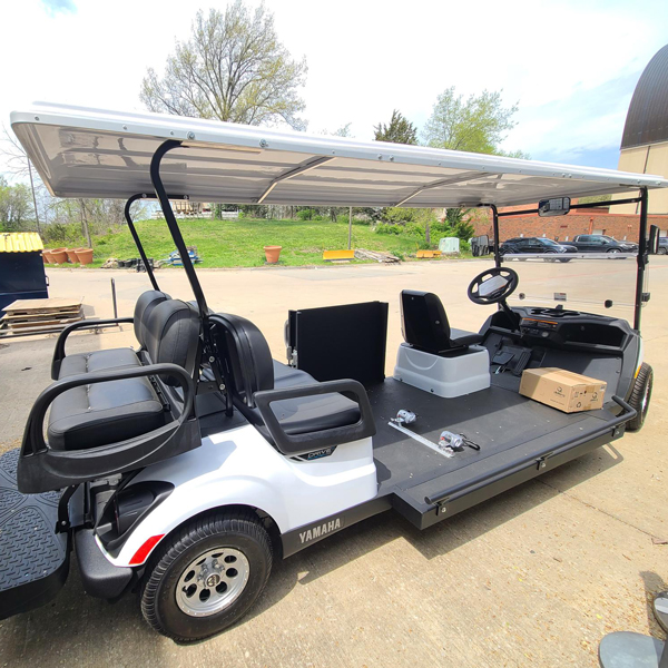 A white Yamaha extended golf cart with black seating and a canopy roof is parked on a paved surface. The cart has three rows of seats and an open central platform, designed for wheelchair accessibility.
