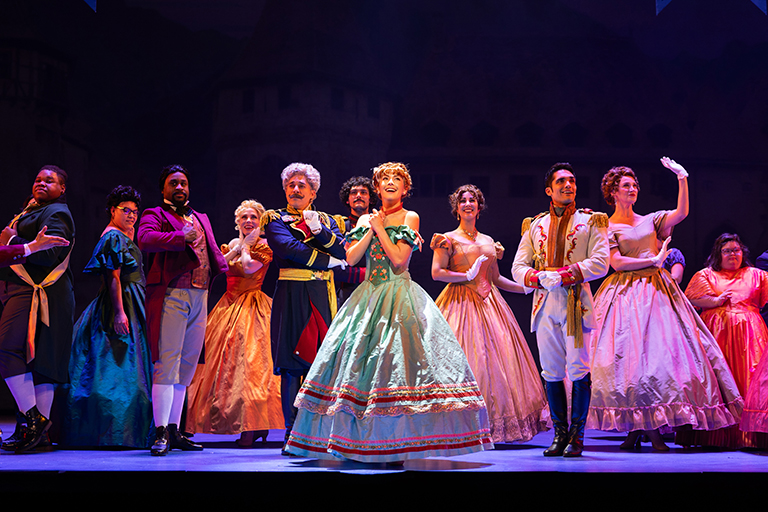 A lively ensemble cast performs a grand ballroom scene from Disney's Frozen on stage. At the center, Anna, dressed in a green and gold gown with floral accents, clasps her hands with excitement. To her right, Hans stands confidently in a white military-style jacket with gold details. Surrounding them, elegantly dressed courtiers in colorful gowns and formal attire strike a regal pose, exuding joy and sophistication. The stage lighting casts a warm glow, highlighting the grandeur of the moment against a backdrop of a grand castle.
