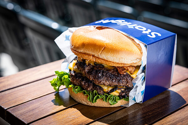 A juicy bacon double cheeseburger with lettuce, melted cheese, and two beef patties, served in a blue box labeled "Starlight," sitting on a wooden table in an outdoor setting.