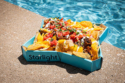 A tray of loaded nachos topped with melted cheese, shredded meat, diced tomatoes, onions, jalapeños, and cilantro, served in a blue star-shaped container labeled "Starlight," sitting on a fountain ledge with clear blue water in the background.