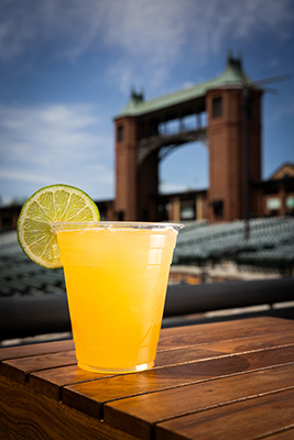 A plastic cup filled with a bright yellow-orange cocktail garnished with a slice of lime sits on a wooden table. In the background, there are stadium-style seats and a large brick structure with towers, likely part of an outdoor theater venue, under a blue sky with light clouds.