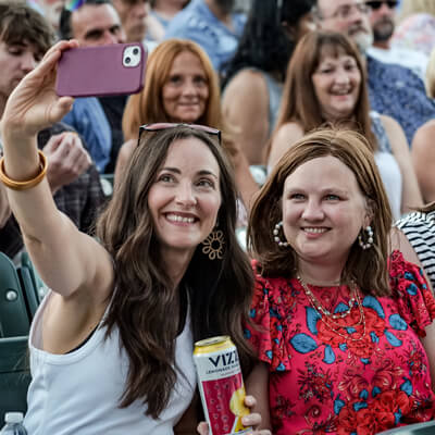 Two women smiling and taking a selfie at an outdoor event, with one holding a can of Vizzy hard seltzer. A crowd of people is seated in the background.
