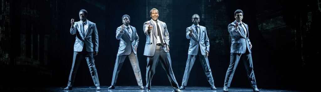 Five performers in matching gray suits sing and dance in unison on a dark stage during a scene from the musical Ain’t Too Proud – The Life and Times of The Temptations, with dramatic lighting highlighting the center performer.