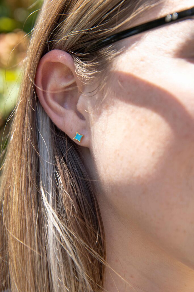 A woman is wearing a stud earring. The earring is blue and looks like a Starlight star. She is standing with trees in the background.