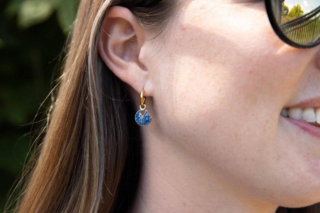 Woman wearing one of the earring styles. The dangly earring is the dark blue starlight logo with a gold hoop. The woman is seen smiling.