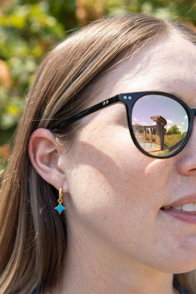 A woman is wearing a pair of dangly  gold earrings. The earrings have a dangly blue Starlight Star attached. The woman is standing in front of trees wearing sunglasses. In the reflection of the sunglasses, the Starlight fountain can be seen.
