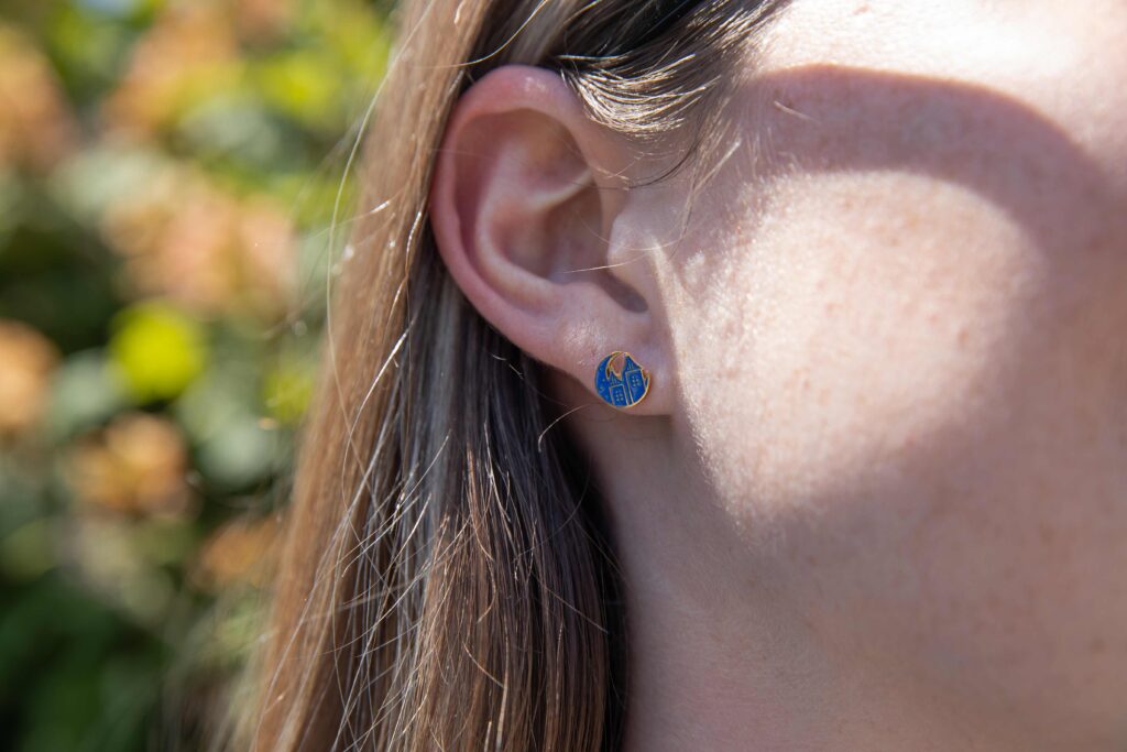 A woman is wearing blue stud earrings. The earrings are the blue Starlight logo. The woman is standing in front of trees in the venue.