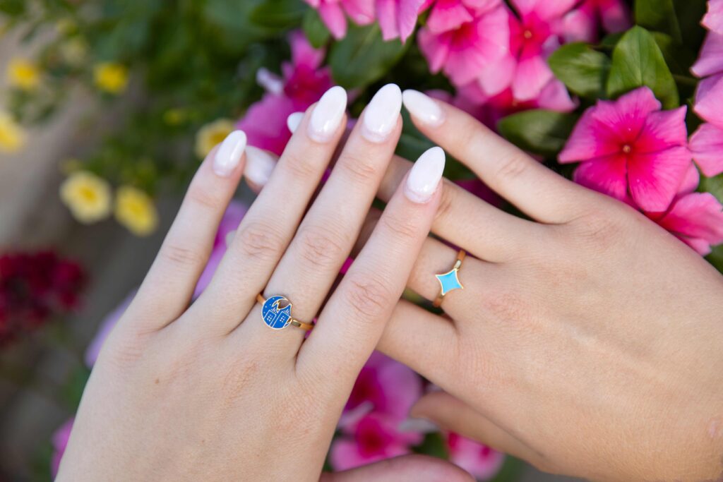2 gold rings with blue starlight gems on 2 hands in front of pink flowers.