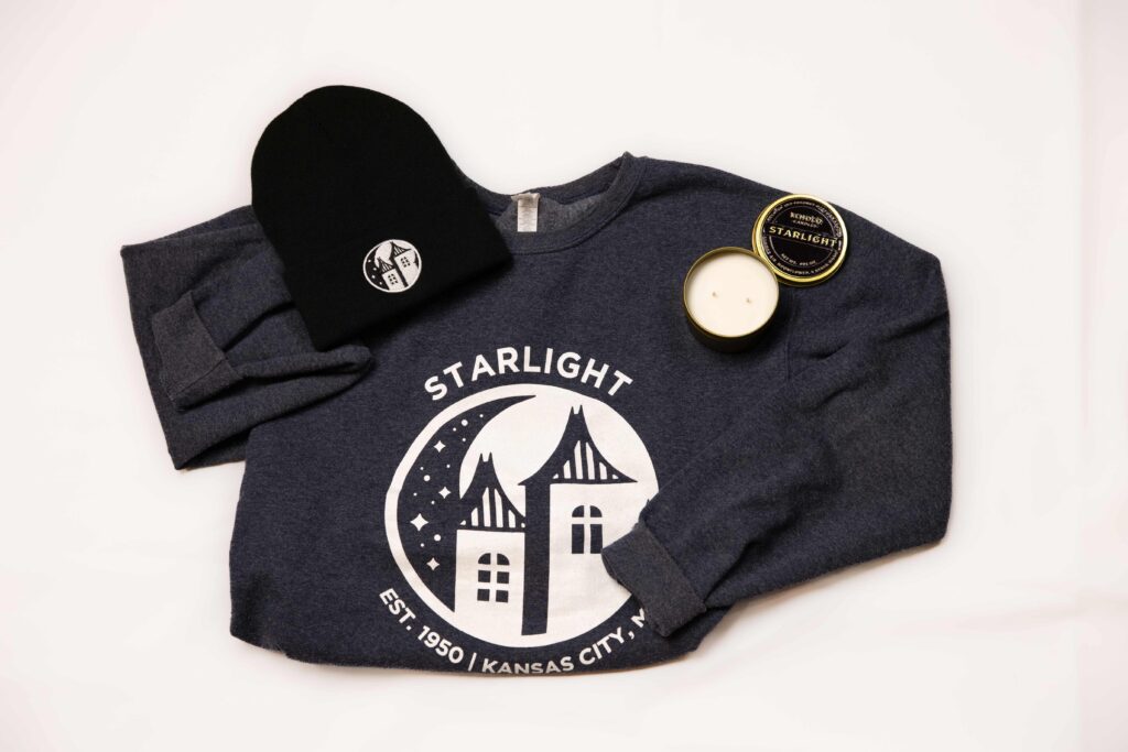A dark gray Starlight Sweatshirt lays on a white background. The sweatshirt has a white Starlight logo with the words "Starlight" and "est. 1950 - Kansas City, MO." Next to the sweatshirt is a Starlight beanie and a gold Starlight candle. 