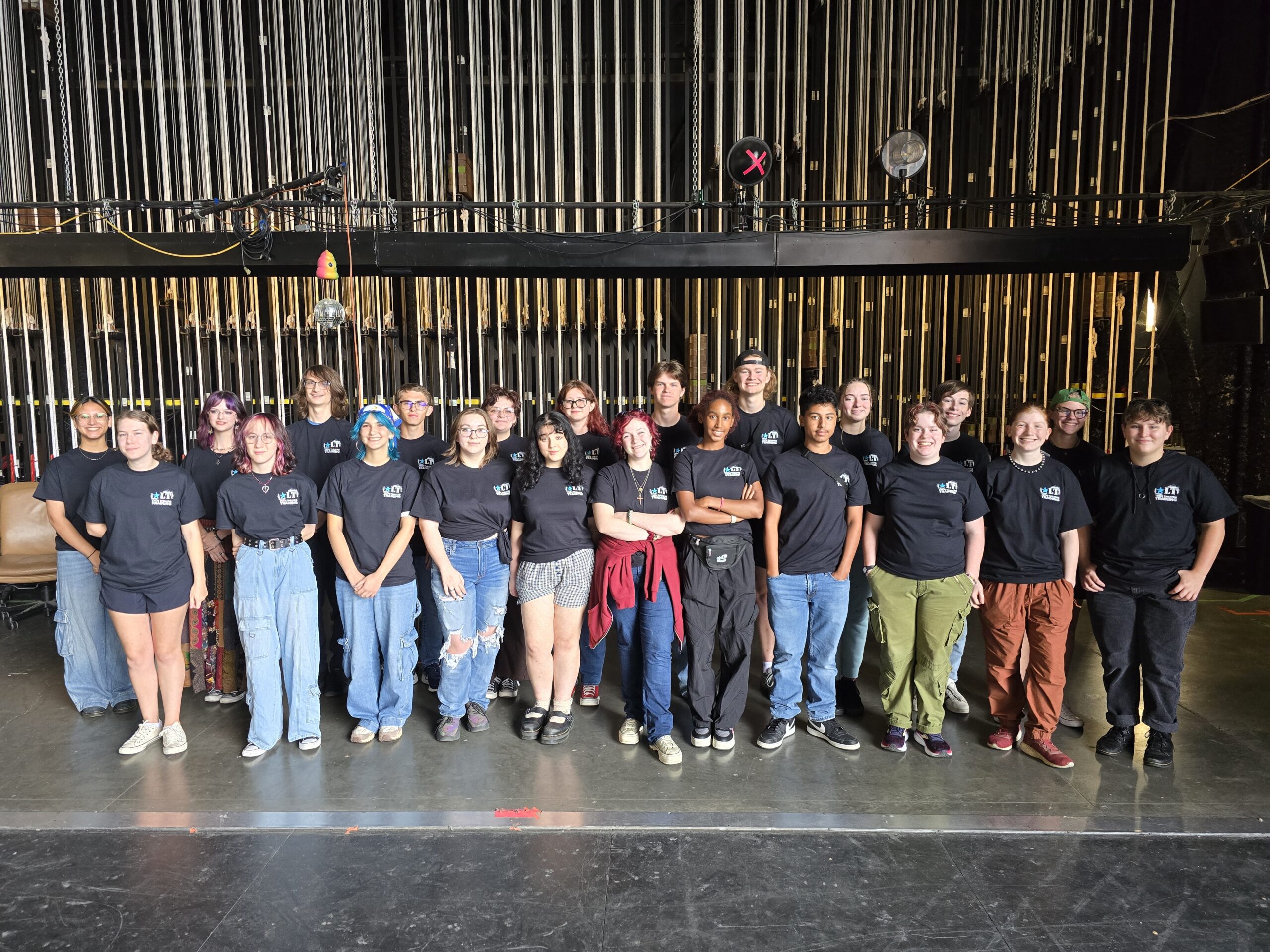A group of 22 students in Tech Theatre Training shirts standing posed for a photo in Starlight's stagehouse.