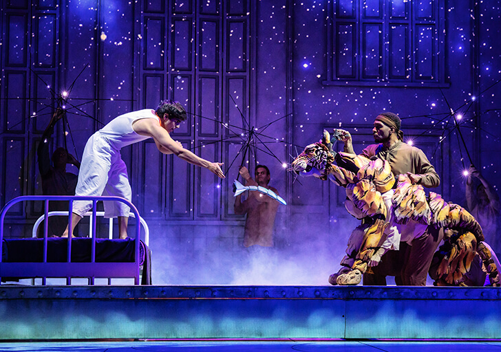 Stage scene from a theatrical production featuring an actor in white reaching from a bed toward a life-sized tiger puppet operated by puppeteers, with a starry blue backdrop.