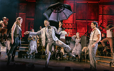 A lively stage performance from the musical Hadestown featuring a performer in a silver suit dancing with an open black umbrella, surrounded by cast members in vintage-style costumes and a warm, dramatic red-and-gold lighting backdrop.