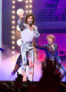 An energetic concert-style scene from the musical A Beautiful Noise featuring a performer in a shimmering silver outfit singing into a microphone as dancers and musicians perform around him under glowing purple and pink stage lights.