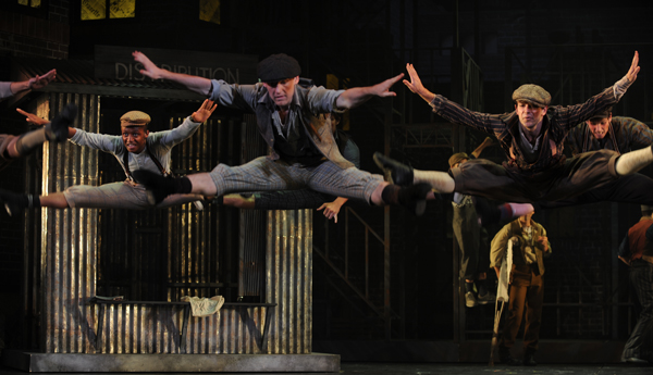 Dancers in the Newsies stage production high in the air doing split jumps.