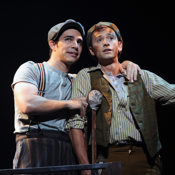 Two actors portraying Jack Kelly and Crutchie in Newsies stand together onstage, wearing period costumes and newsboy caps.