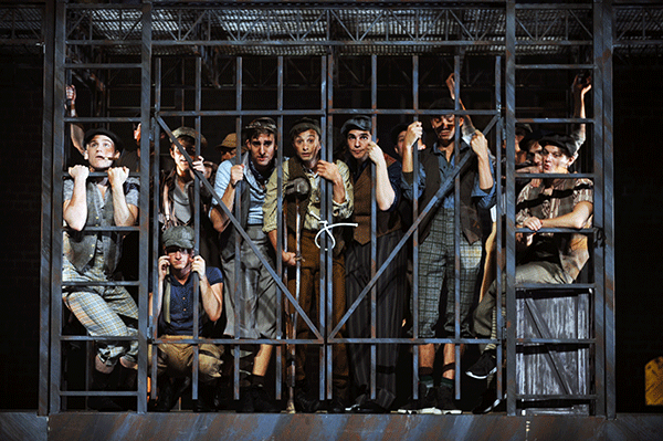 A group of newsies stand behind bars in a jail cell during a production of Newsies.