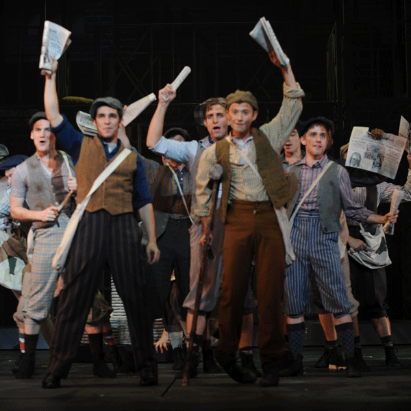 Cast dressed as newspaper boys hold up newspapers during a production of Newsies.