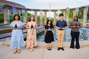 Five students stand outdoors beside a reflecting pool, holding awards and smiling for a group photo. They are dressed in semi-formal attire, with a modern brick-and-wood structure and landscaped grounds in the background.