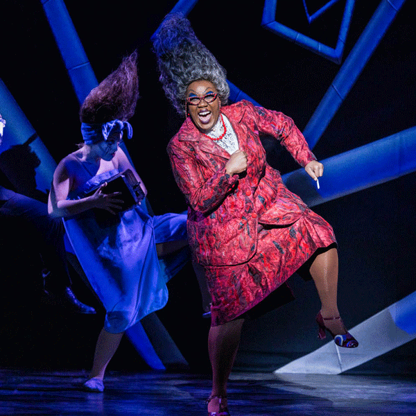 A lively musical number from the musical Beetlejuice featuring a performer in a bright red patterned suit and towering gray wig dancing energetically among quirky costumed cast members.