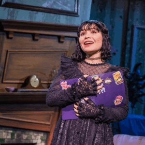 Lydia is dressed in black goth clothing and holds a book as she sings.