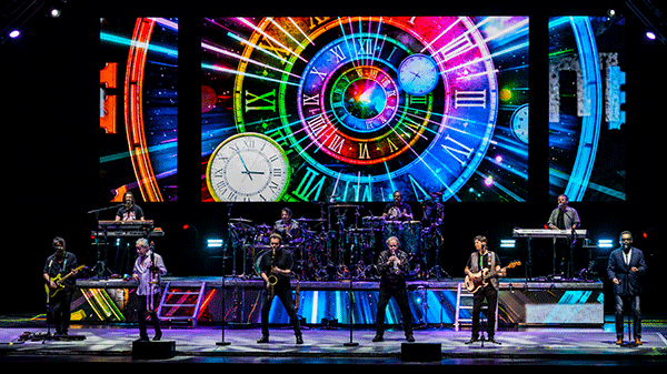 The band Chicago performs on stage with bright colorful images of clocks behind them.
