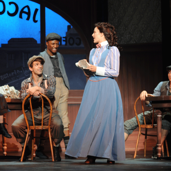 A woman dressed in late 19th-century clothing stands while talking to Newsies.