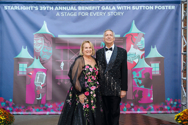 A couple in a tuxedo and gown stand in front of a back drop at the Starlight Gala.
