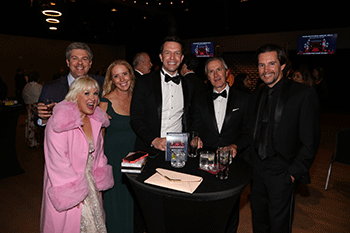 A group of people in formal wear enjoy drinks around a table.