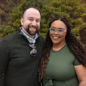 A white man with dark hair and a beard wearing a button down shirt with a sweater and a black woman with long dark hair wearing a green dress