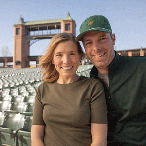 A white woman with light brown hair wearing a dark green shirt and a white man wearing a green baseball hat and shirt