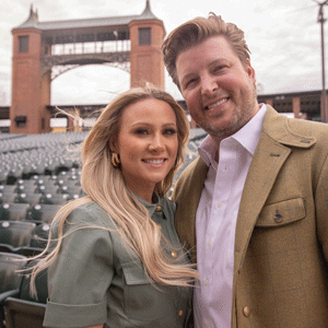A white woman with long blond hair wearing a green shirt and a white man with light brown hair wearing a white shirt with a brown jacket