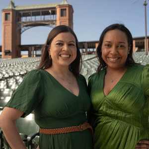A white woman with long brown hair wearing a green dress and a black woman with dark hair wearing an emerald dress