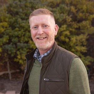 A white man with brown hair and white facial hair wearing a green sweater and a brown vest