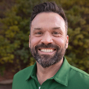 A white man with dark hair and a beard wearing a green button down shirt