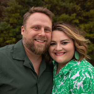 White male with a beard in a dark green shirt and white female with dark blonde hair in a green dress with a floral print