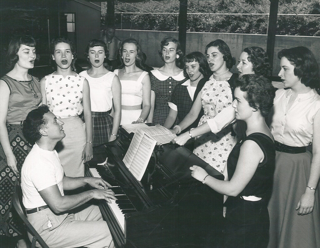 Black and white photo of singers rehearsing with a pianist